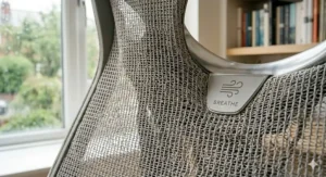 A photorealistic, high-detail macro photograph capturing the complex, open weave structure of the breathable light grey mesh material on the backrest of the ergonomic office chair, with natural light and a soft focus of the rainy UK background.